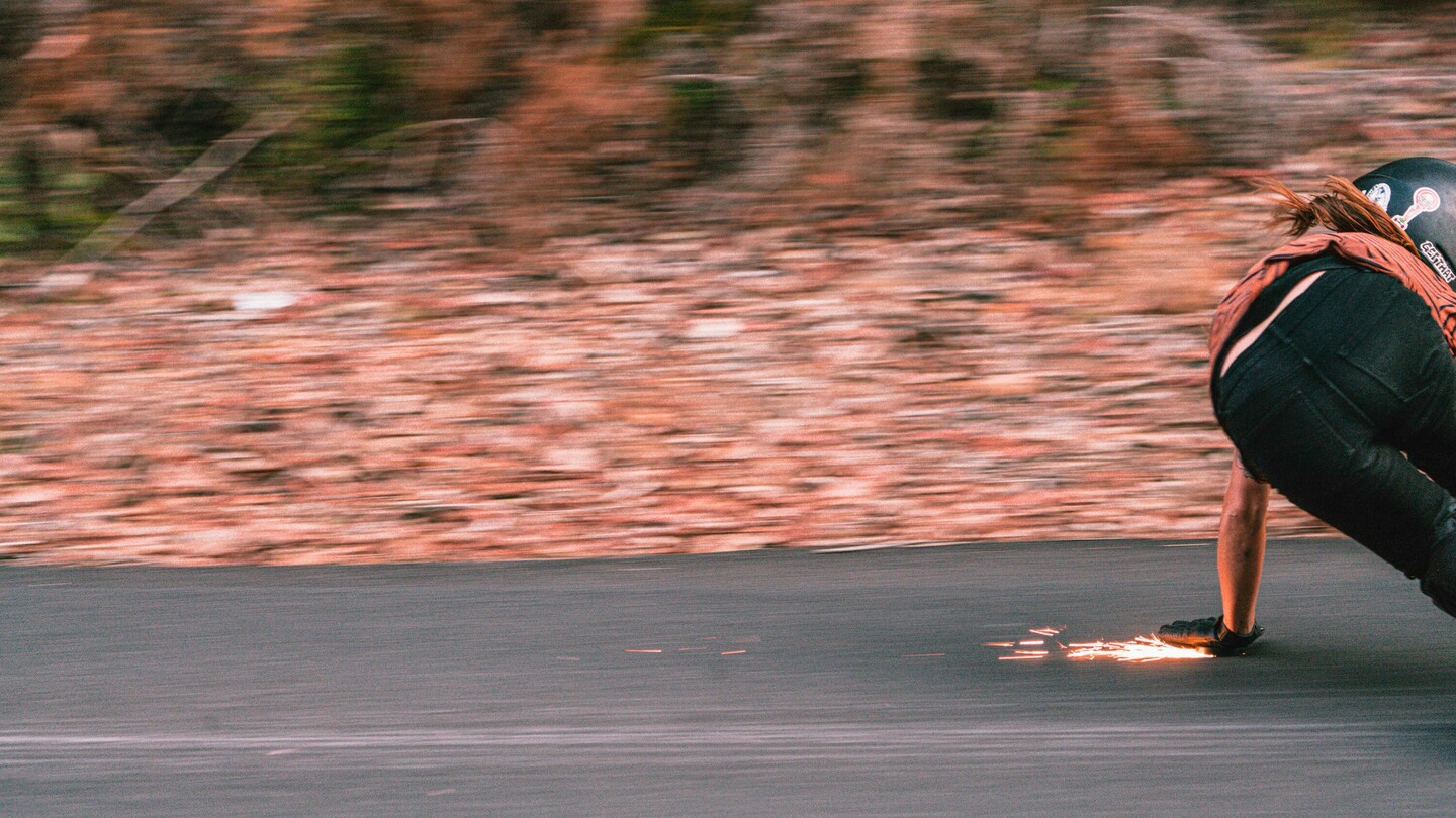 Austrian downhill skateboarder Anna Pixner is captured speeding on her longboard. In a low crouch, she touches the asphalt with her gloved hand, creating sparks. The motion blur in the background highlights the high speed and dynamic nature of the scene as Anna navigates a curve on the road.