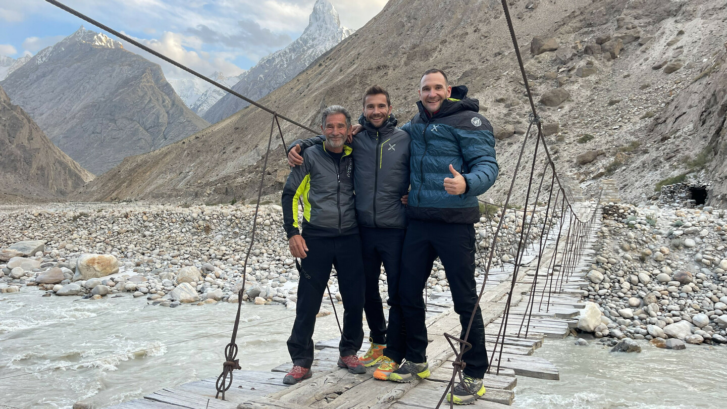 Three climbers (Eduard and Alex Marin with their father) standing and smiling on a suspension bridge over a mountain river, surrounded by rocky terrain and snow-capped peaks in the background.