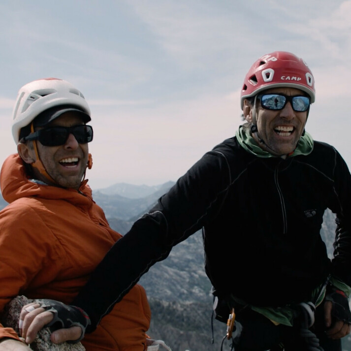 Links im Bild steht Timmy O’Neill in einer orangefarbenen Jacke und einem Helm, lächelnd mit Sonnenbrille. Rechts neben ihm steht der blinde Kletterer Erik Weihenmayer in einer schwarzen Jacke und einem roten Helm. Beide Männer stehen auf einem Berggipfel, wirken glücklich und genießen die weite Berglandschaft im Hintergrund. Das Bild zeigt das erfolgreiche Besteigen eines Gipfels und symbolisiert Abenteuer, Klettern und Motivation.