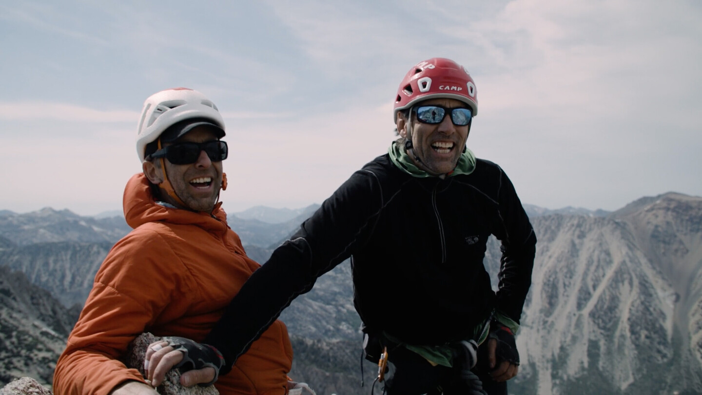 Links im Bild steht Timmy O’Neill in einer orangefarbenen Jacke und einem Helm, lächelnd mit Sonnenbrille. Rechts neben ihm steht der blinde Kletterer Erik Weihenmayer in einer schwarzen Jacke und einem roten Helm. Beide Männer stehen auf einem Berggipfel, wirken glücklich und genießen die weite Berglandschaft im Hintergrund. Das Bild zeigt das erfolgreiche Besteigen eines Gipfels und symbolisiert Abenteuer, Klettern und Motivation.
