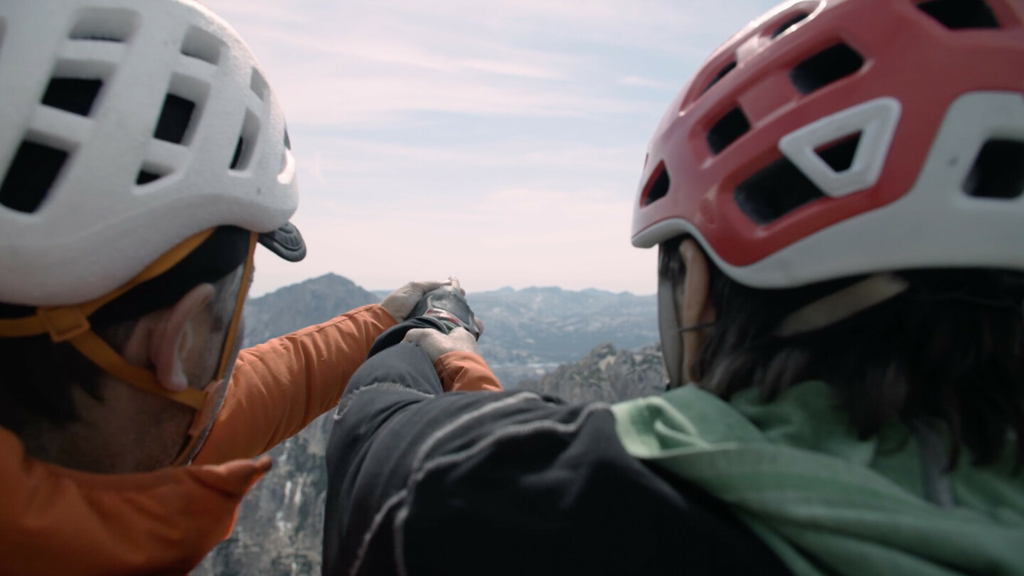 Nahaufnahme von Timmy O’Neill (links) und Erik Weihenmayer (rechts) mit Helmen, während Timmy Eriks Hand führt, um ihm die Berglandschaft zu zeigen. Die Szene verdeutlicht den Teamgeist und die unterstützende Zusammenarbeit der beiden Kletterer, während die Weite der umliegenden Gipfel im Hintergrund zu sehen ist.