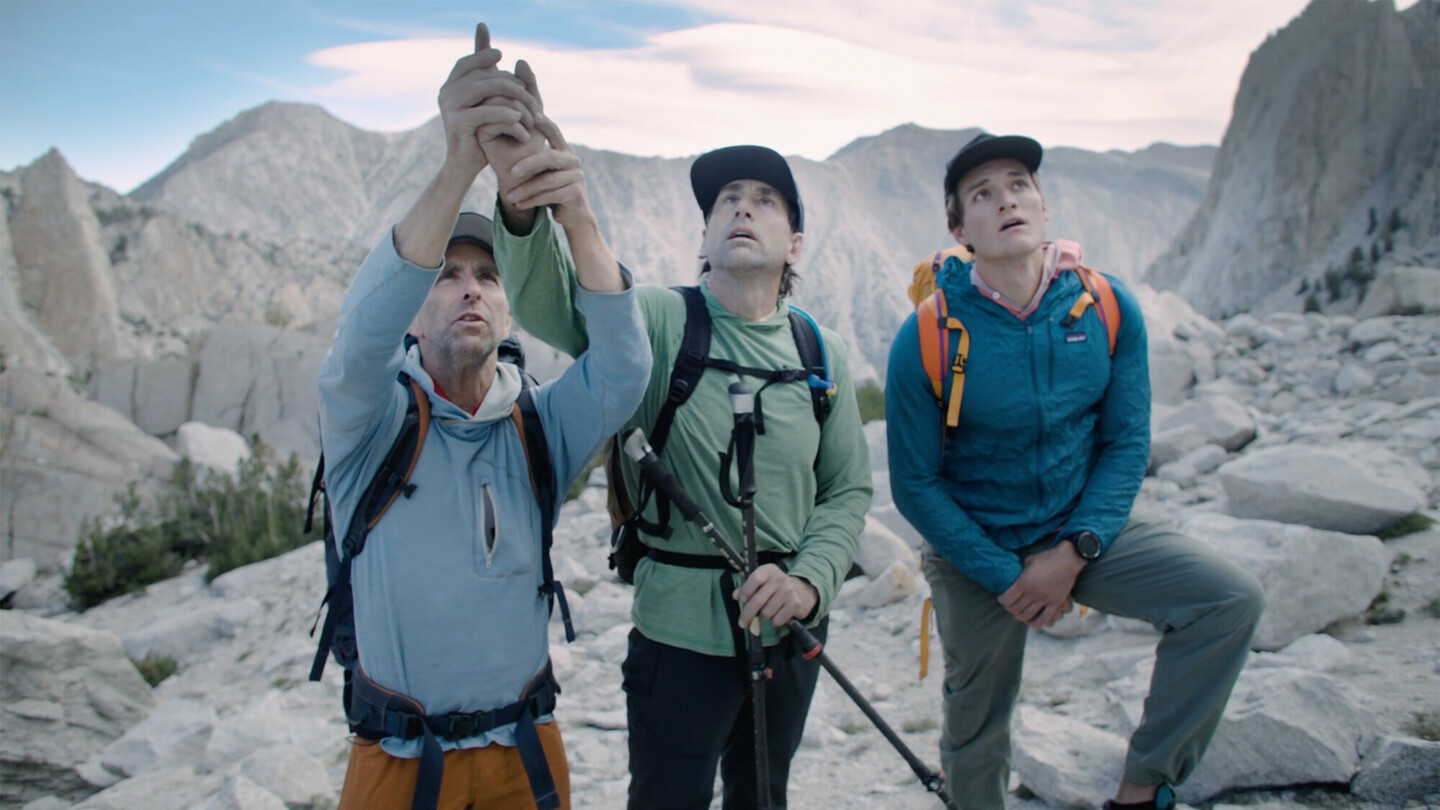 Drei Bergsteiger in Outdoor-Kleidung und mit Rucksäcken stehen in einer felsigen Berglandschaft. Der linke Mann führt die Hand des blinden Mannes in der Mitte, um ihm die Kletterroute und die umliegenden Berge zu zeigen.